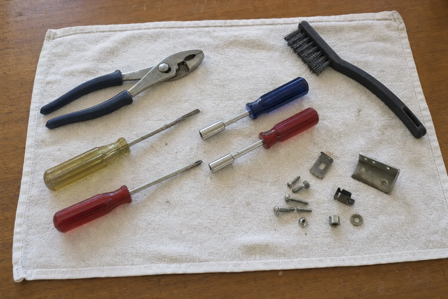 Slip-joint pliers, screwdrivers, nut drivers and a wire brush laid out on a towel beside screws and brackets